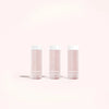 Three Solawave Skin Therapy Activating Serum Refill bottles stand on a light pink background. Transparent with white caps, revealing the pink liquid inside and shadows beneath, they perfectly complement the Solawave Wand treatment and its anti-aging ingredients.