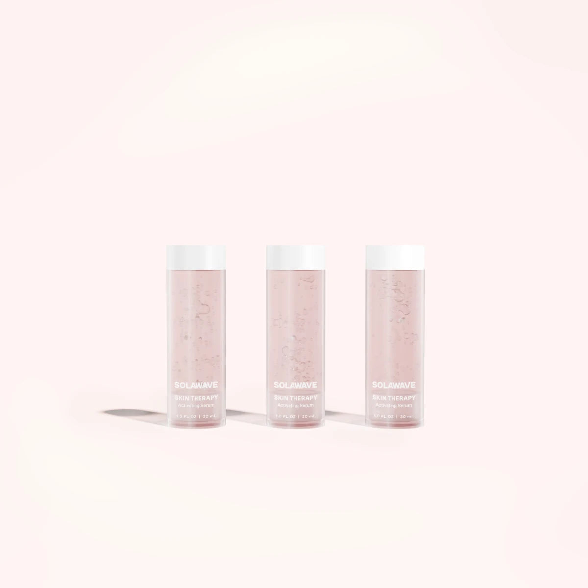 Three Solawave Skin Therapy Activating Serum Refill bottles stand on a light pink background. Transparent with white caps, revealing the pink liquid inside and shadows beneath, they perfectly complement the Solawave Wand treatment and its anti-aging ingredients.